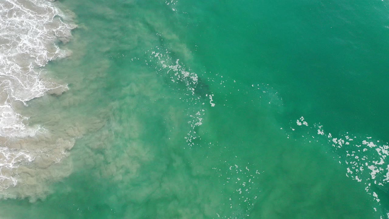 Overhead view of turquoise ocean in Los Cabos, Mexico, with foamy surf at the upper left and scattered bubbles across open water, no visible people.