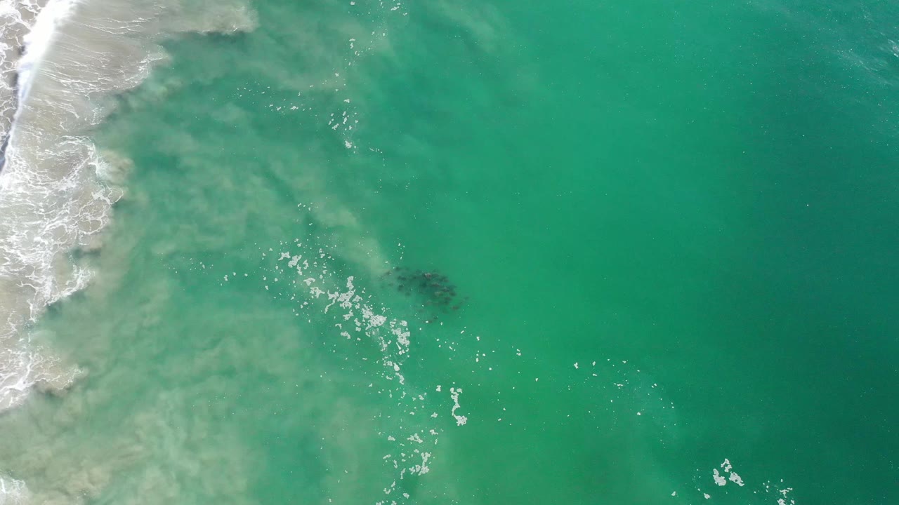Overhead Los Cabos water frame showing turquoise sea, pale churned surf along the left side, a cluster of white foam specks, a dark shape under the surface to the right, and no visible people.
