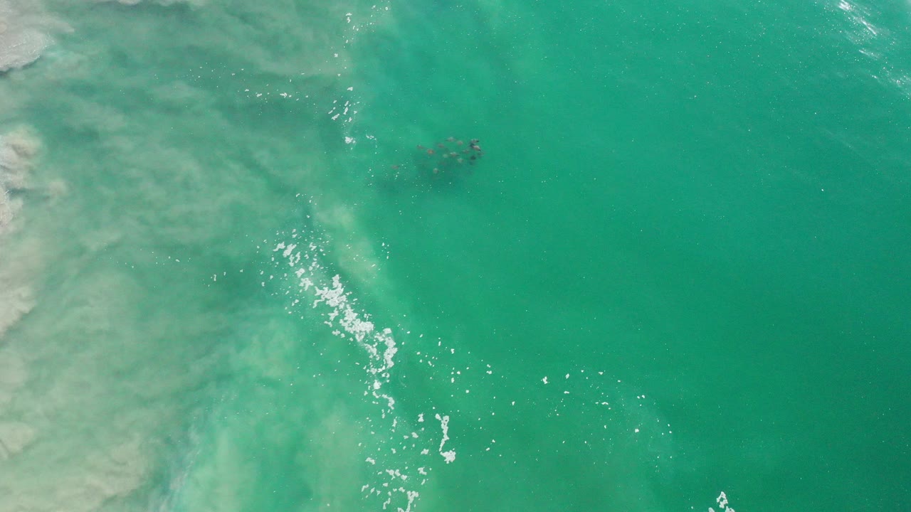 Overhead view of turquoise water in Los Cabos, Mexico, with pale wave foam along the left side, sun glints near center, and no visible people.
