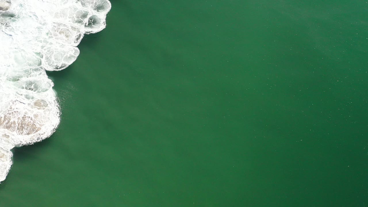 Top-down aerial view in Los Cabos, Mexico with green ocean filling most of the frame, white breaking surf along the left edge, and no visible people.