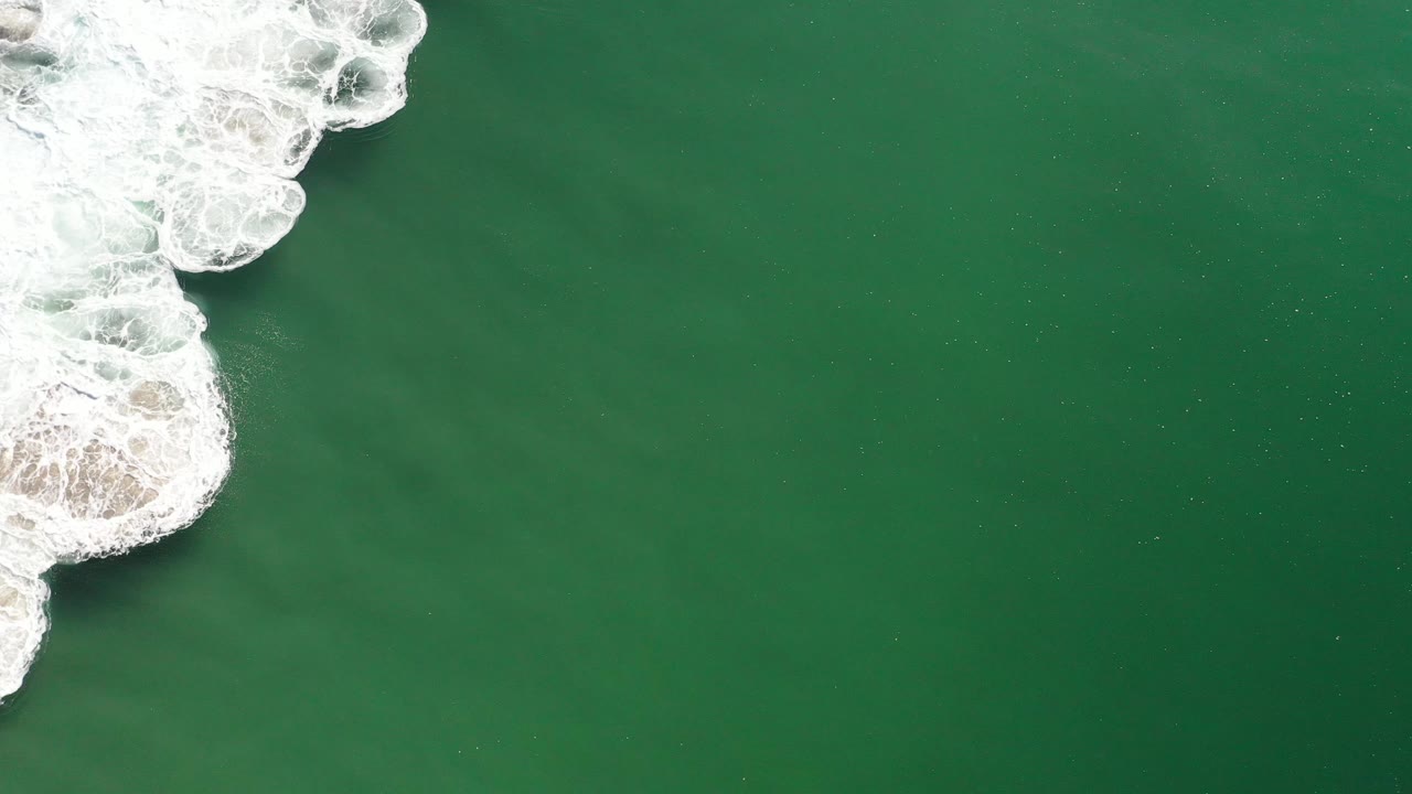 Aerial frame over Los Cabos, Mexico showing a curled band of white surf and pale sand at left against uninterrupted green water, with no visible people.
