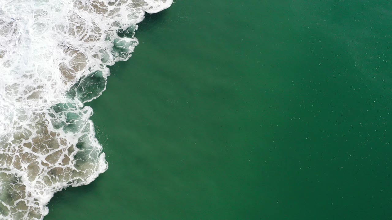 Top-down view of green ocean water in Los Cabos, Mexico, with white foaming surf along the left edge and no visible people.