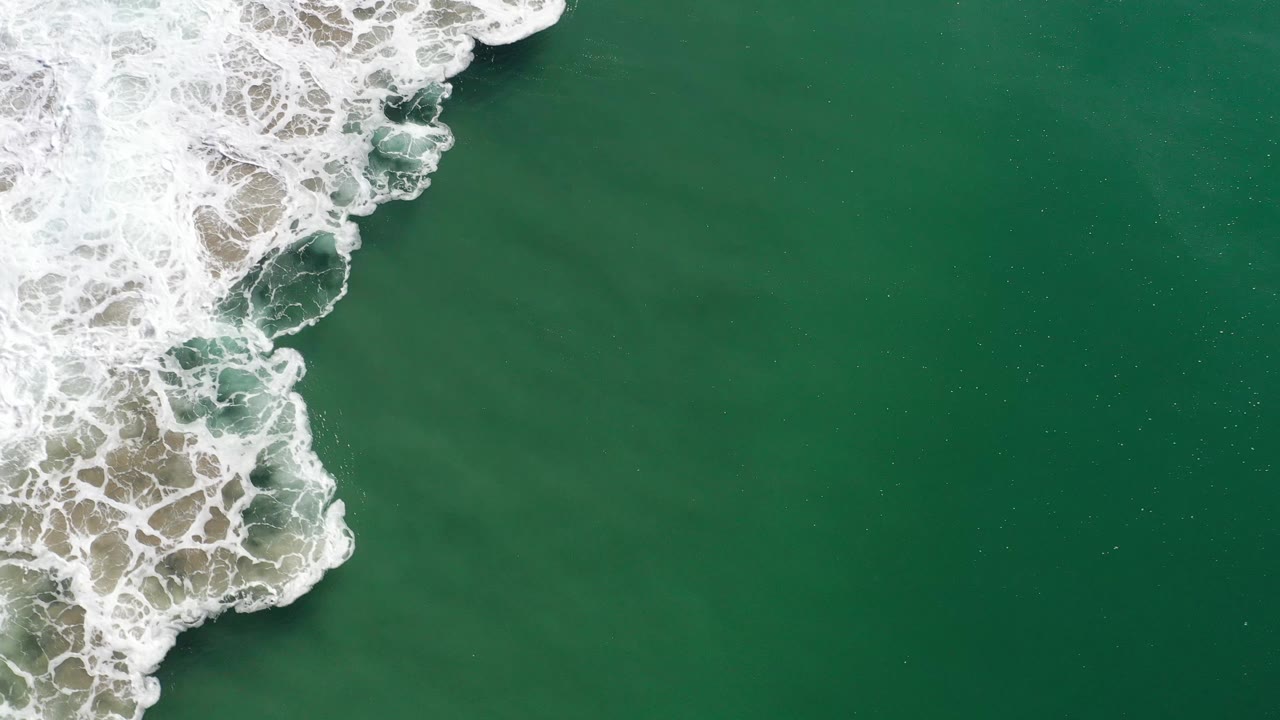 Aerial frame over Los Cabos green ocean as white wave foam pushes farther down the left side, with open water to the right and no visible people.