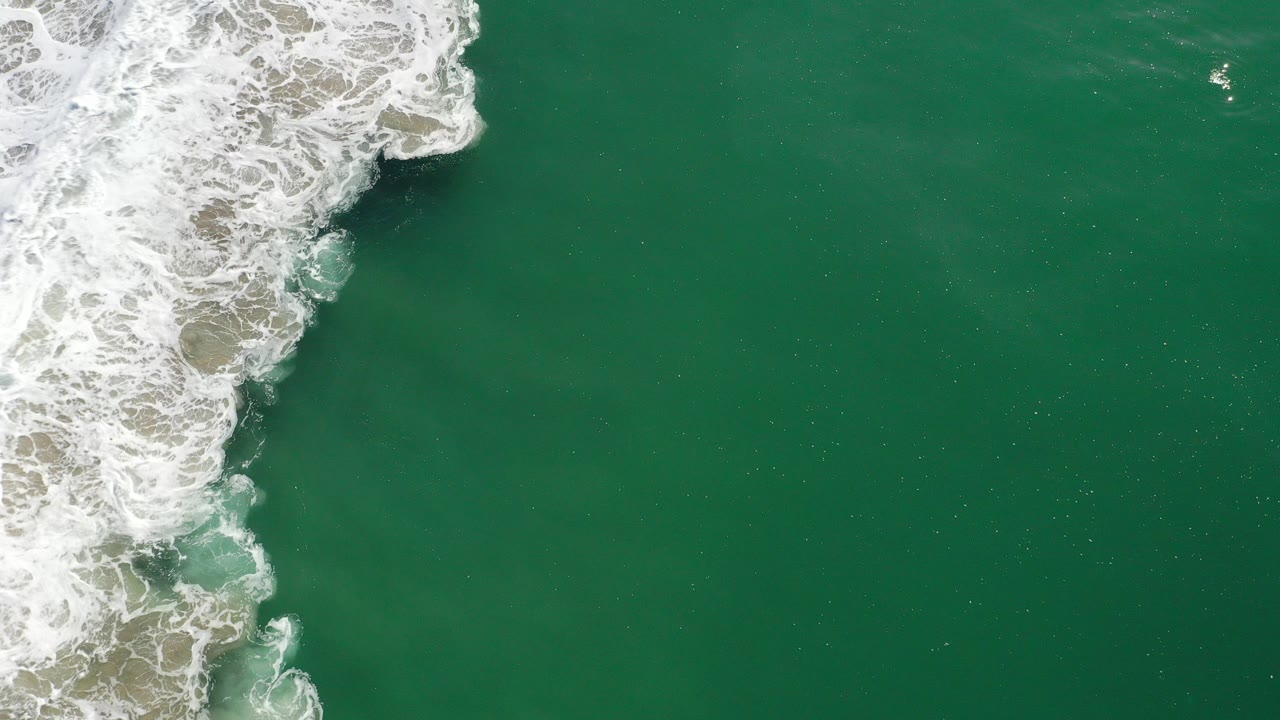 Overhead view in Los Cabos, Mexico, with foamy white surf pushing in from the upper left over teal-green water and no visible people.