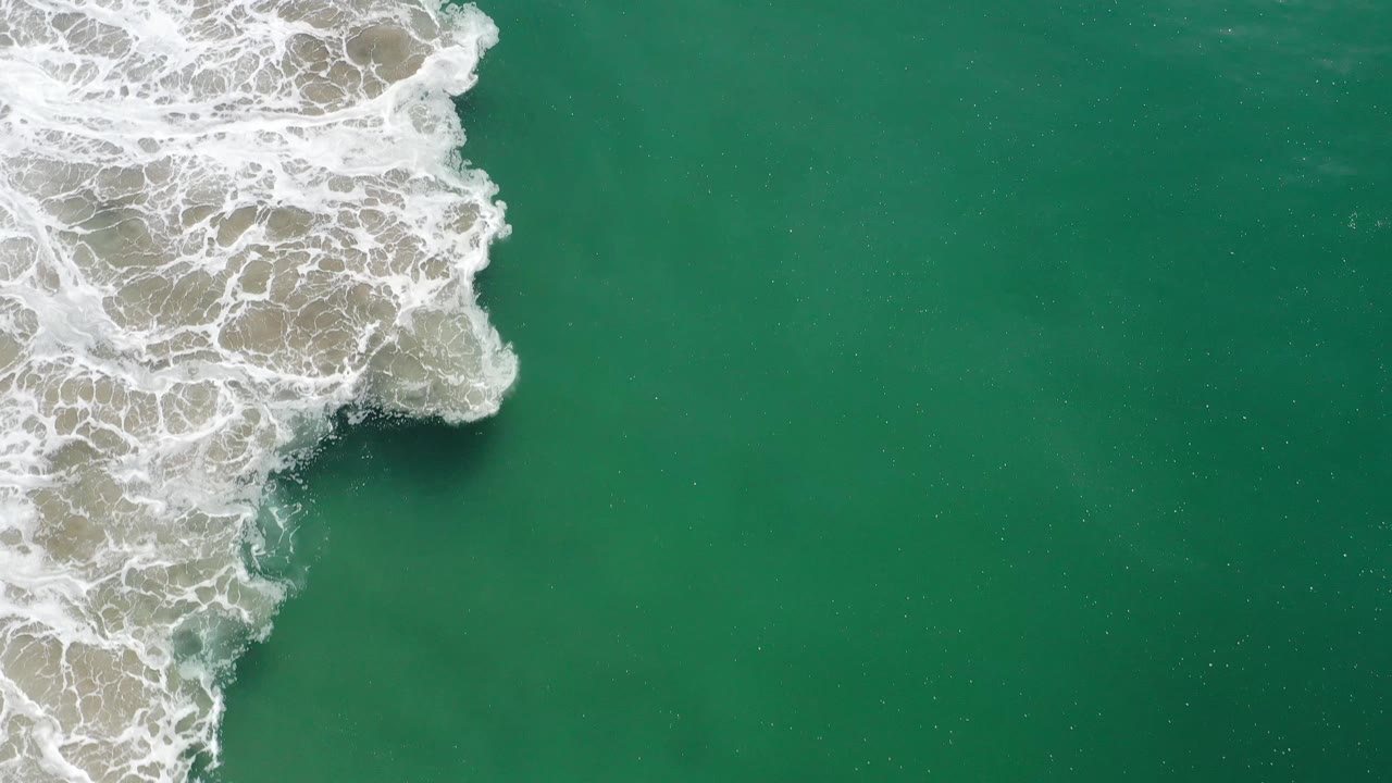 Overhead Los Cabos, Mexico frame with the whitewater shifted down the left side and a broad field of green water to the right, with no visible people.