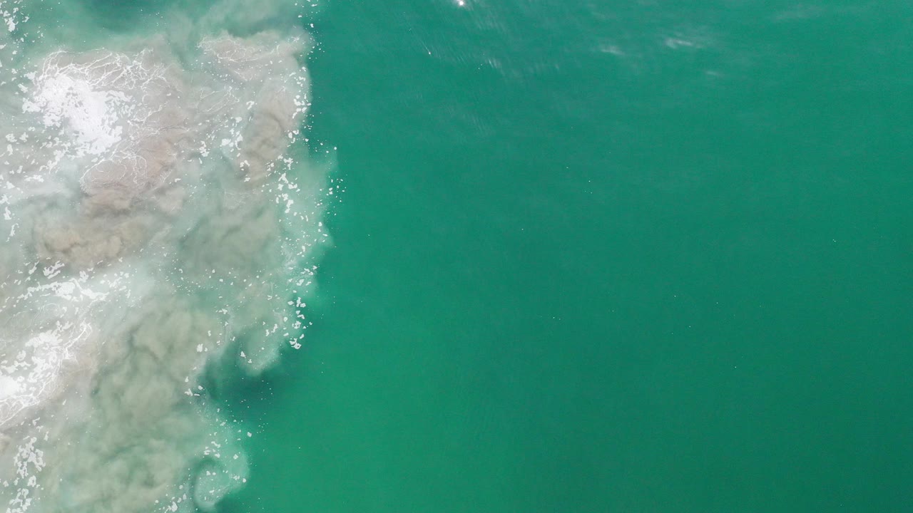 Top-down view of green ocean water in Los Cabos, Mexico, with pale sand or foam patterns entering from the upper left and no visible people.