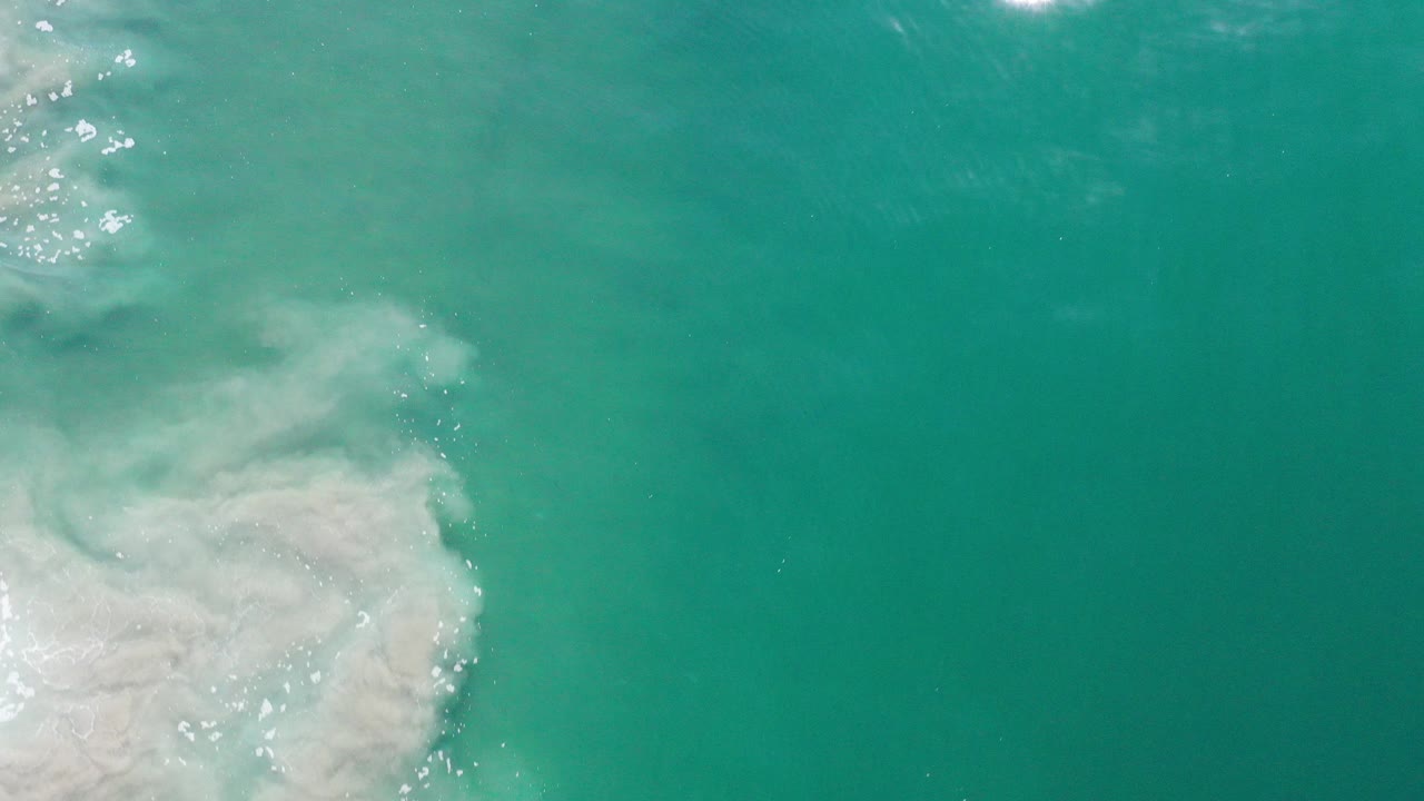 Overhead view of turquoise ocean in Los Cabos, Mexico, with a pale foamy wave mass entering from the lower left and no visible people.