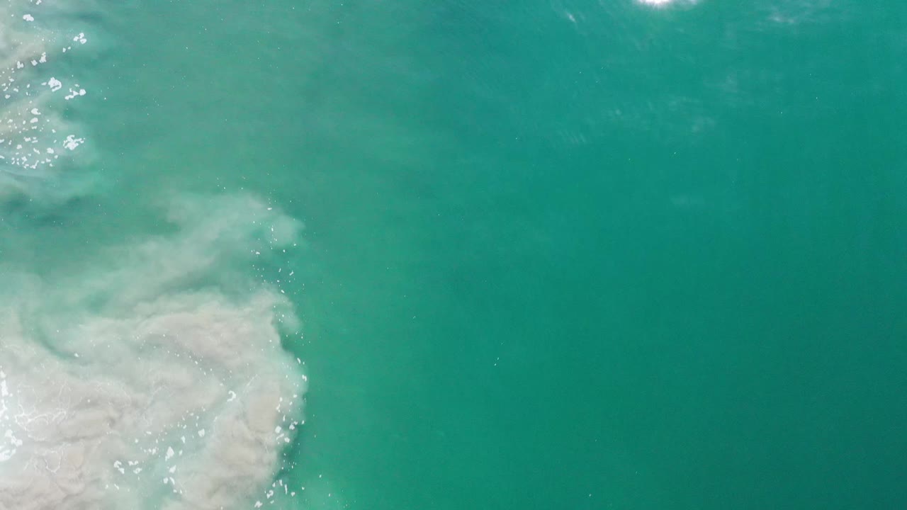 Overhead Los Cabos ocean frame with white foam spreading from the left edge into open teal water and no visible people.