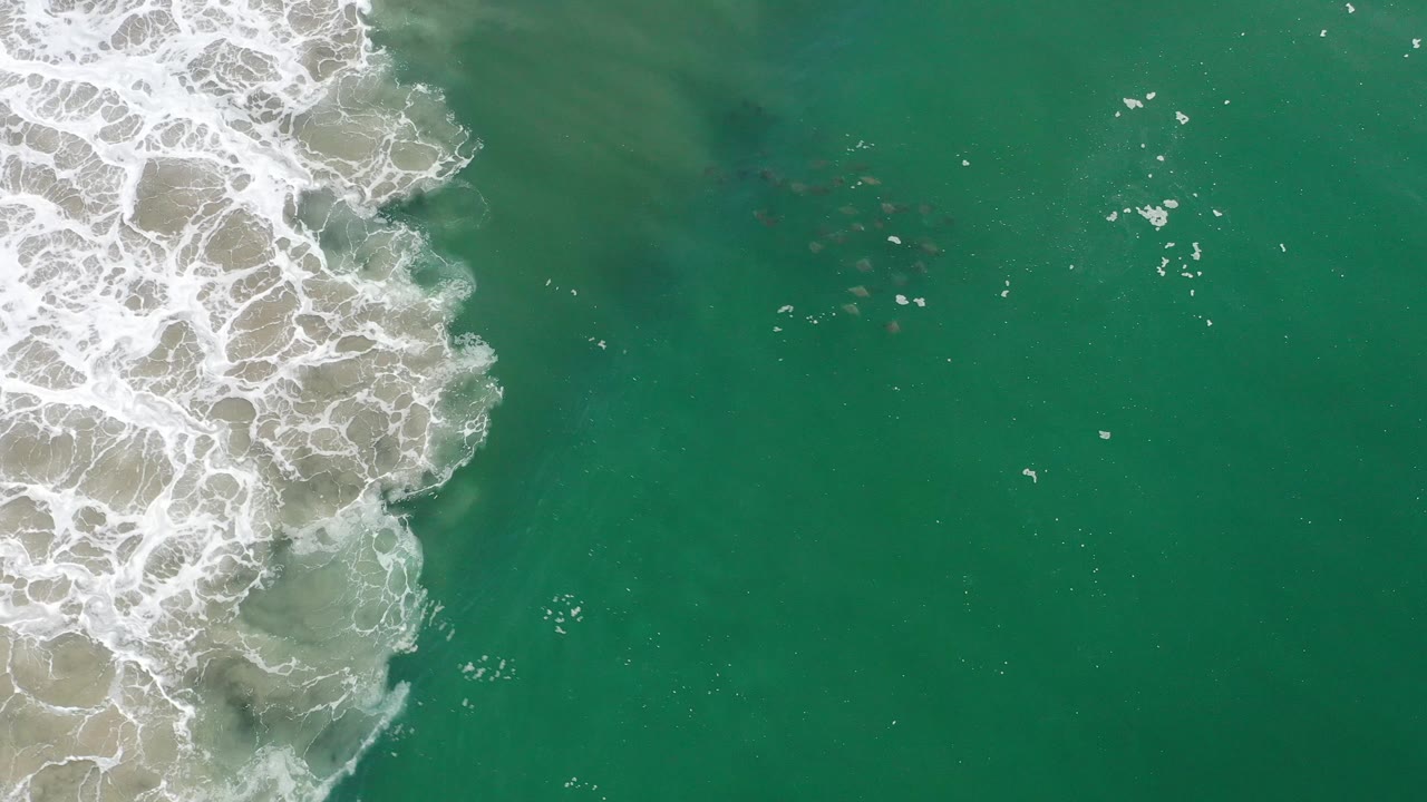 Top-down Los Cabos shot with textured white surf and sandy wash on the left, green ocean on the right, and no visible people or boats.