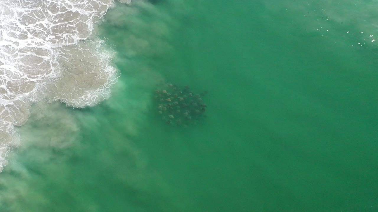 Overhead frame from Los Cabos, Mexico showing green ocean water, white surf foam at the upper left, pale sand beneath shallow water, and a dark submerged rock cluster near center, with no visible people.
