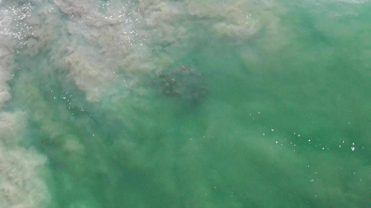 Green coastal water in Los Cabos, Mexico, fills the frame as pale cloudy swirls sit near the top and tiny bright reflections cross the midframe, with no visible people.