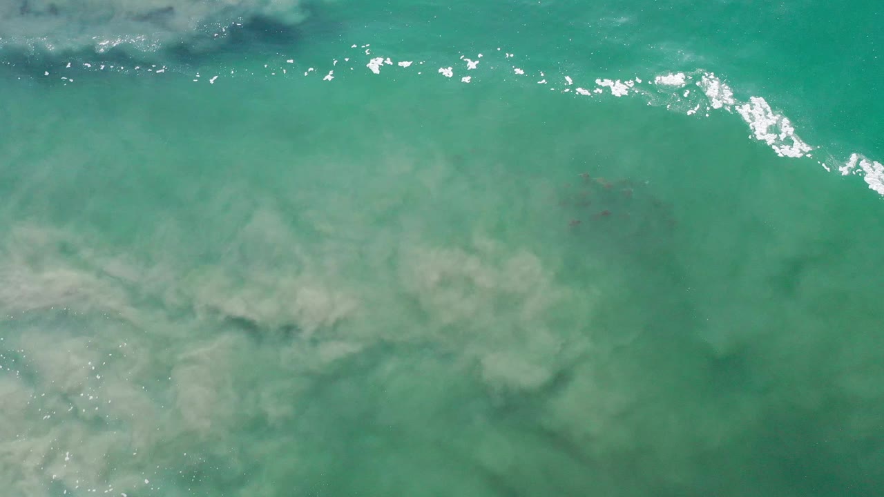 A white bubble wave crosses the upper area of green Los Cabos seawater while muted sand clouds spread underneath, no visible people.