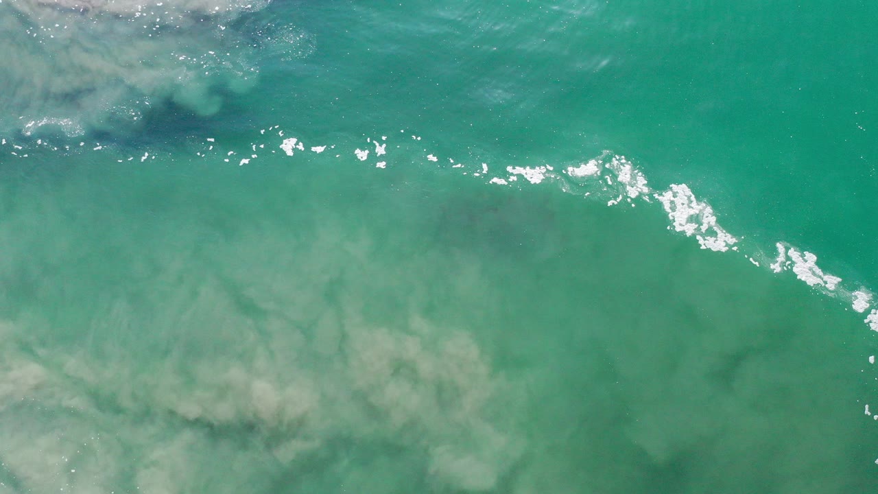 Overhead view of sea-green water in Los Cabos, Mexico, with a diagonal streak of white foam and sun reflections near the surface, no visible people.