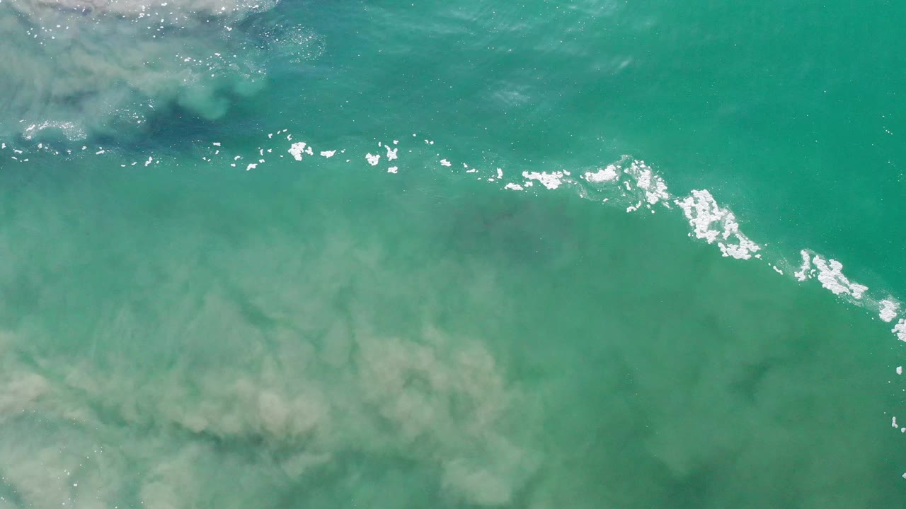 Aerial-looking frame of turquoise Los Cabos water with pale underwater patches, scattered white glints, and a small foamy area at the upper left, no visible people.