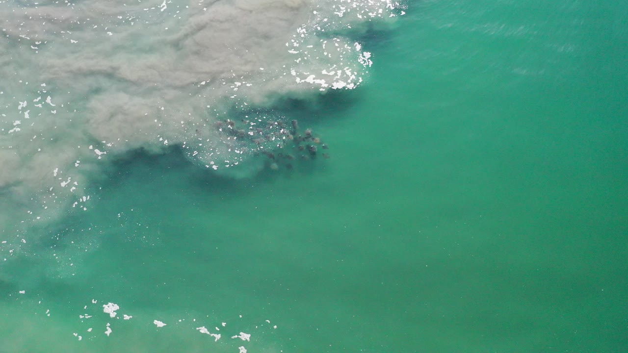 Overhead view in Los Cabos, Mexico of a dark school of fish beneath turquoise water near a band of white sea foam, with no visible people.