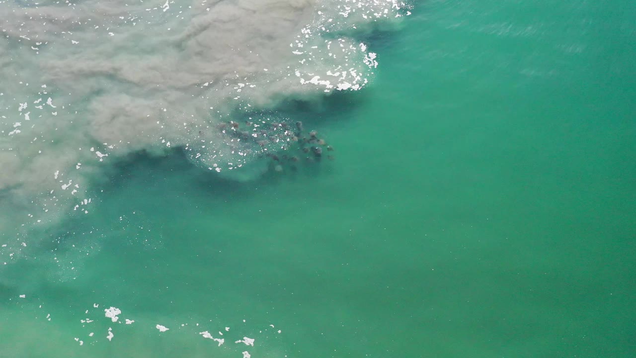 Overhead Los Cabos ocean frame with the fish cluster partly shadowed below pale surf foam and green-blue water, with no visible people.
