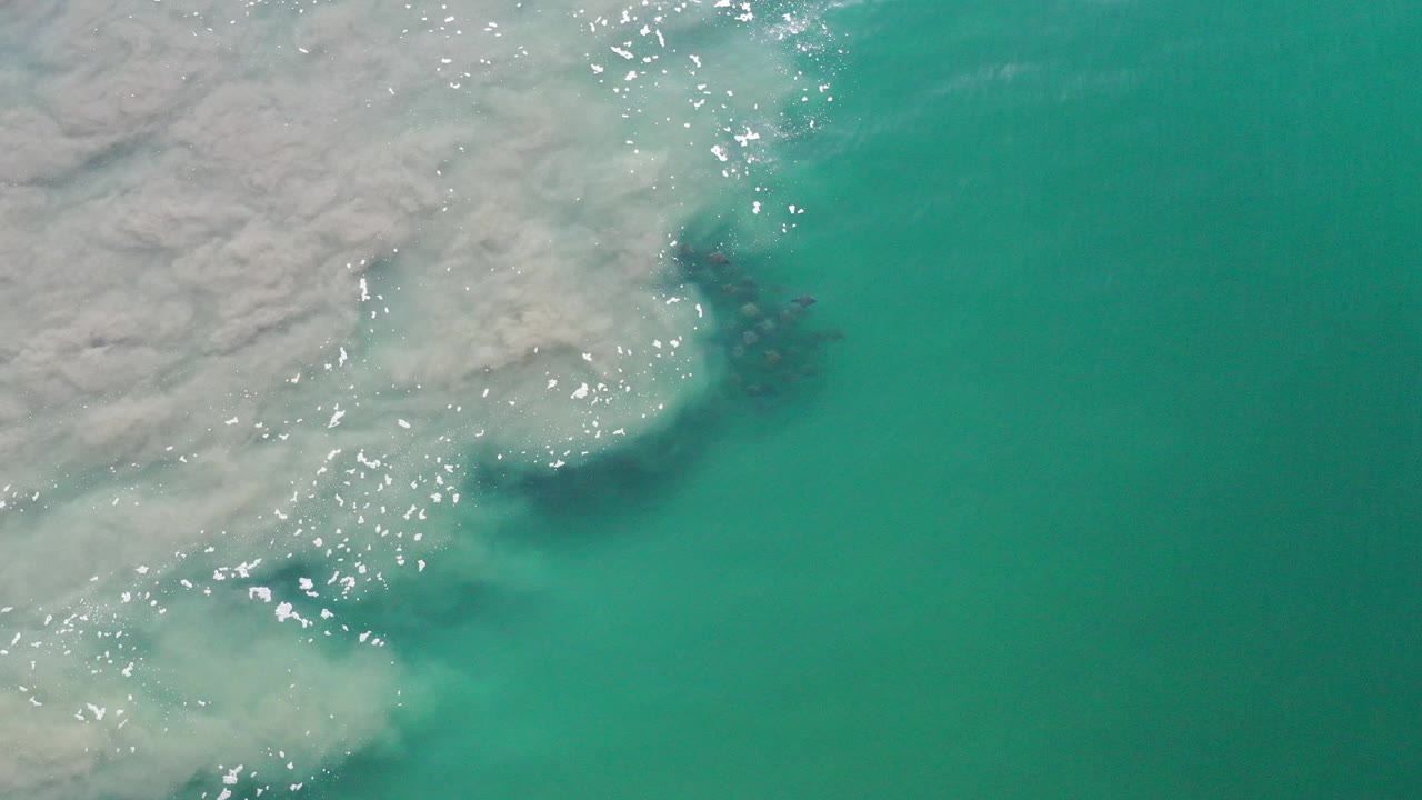 Opening overhead view of Los Cabos teal water meeting pale shoreline foam, with bright surface glints and no visible people.