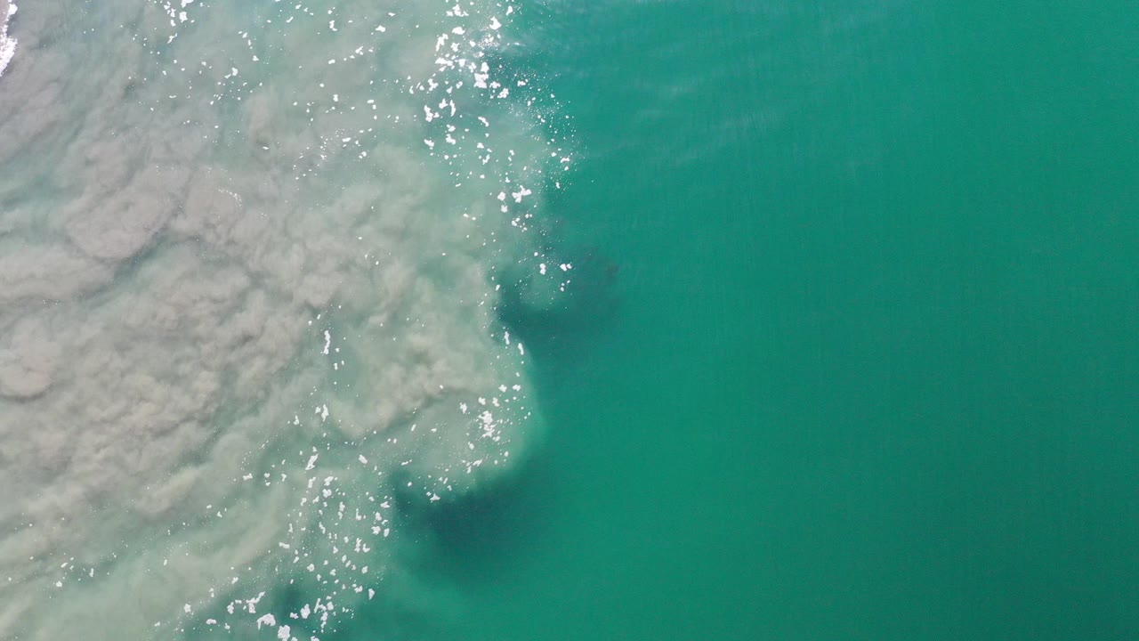 Overhead view of turquoise ocean in Los Cabos, Mexico, with pale foam spreading from the left edge, bright water reflections, and no visible people.