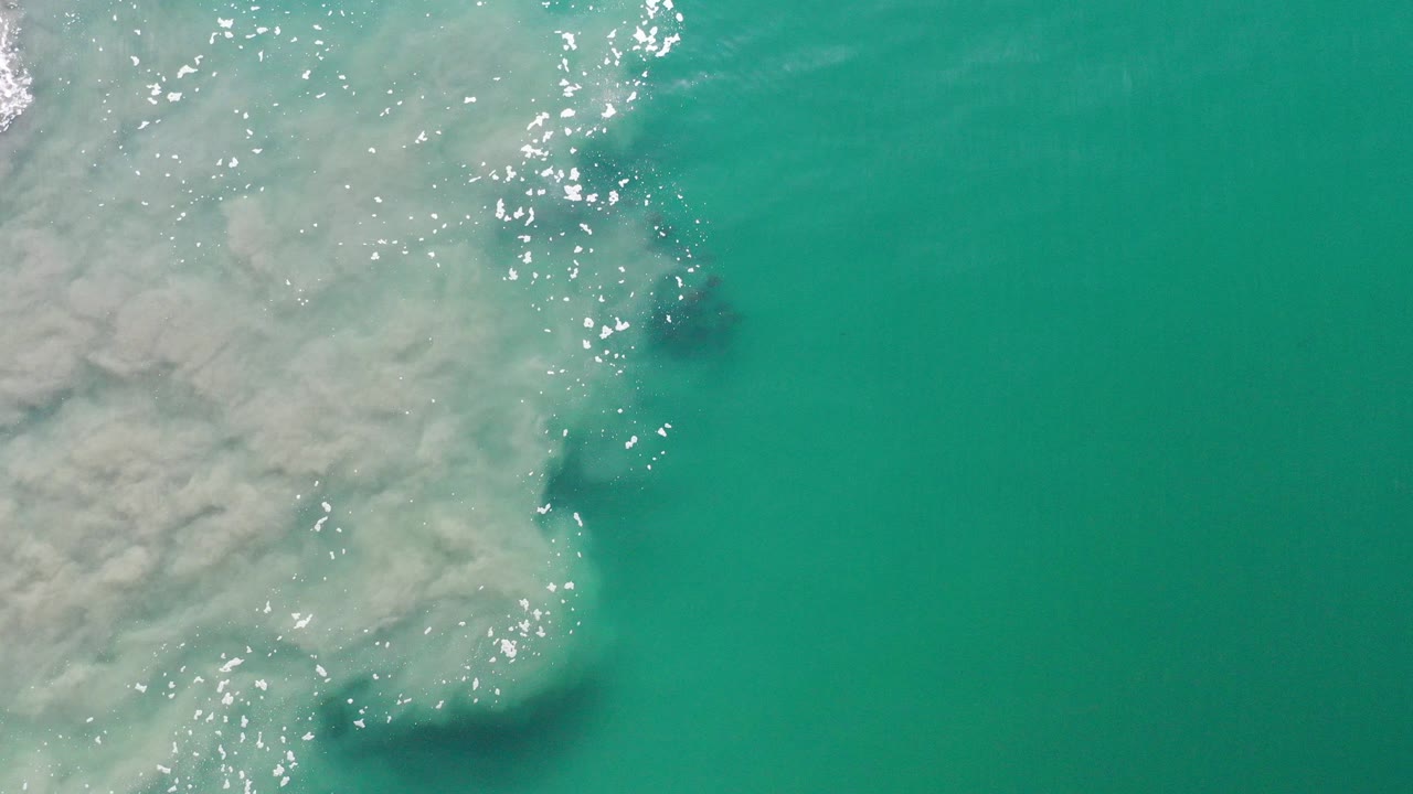 Overhead view of teal seawater in Los Cabos, Mexico, with pale underwater sediment filling the left side, darker patches below the surface, bright sun glints, and no visible people.
