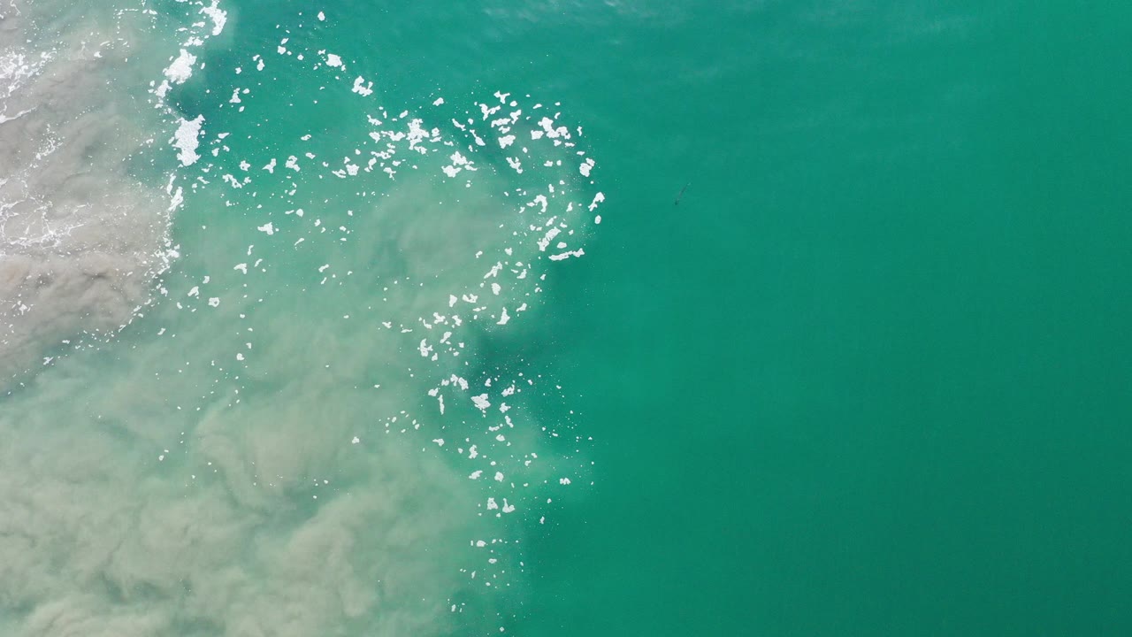 Overhead view in Los Cabos, Mexico, with turquoise water covering most of the frame and pale sand or sediment entering from the left, no visible people.