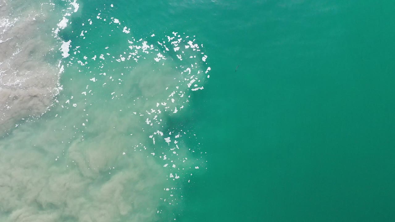 Overhead Los Cabos water detail with a larger patch of pale sandy shallows on the left and scattered white sun reflections across the center, no visible people.