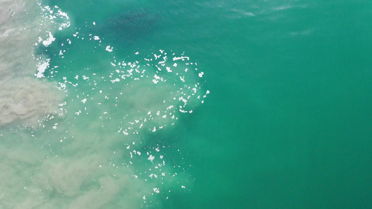 Overhead view of green-turquoise water in Los Cabos, Mexico, with pale foam entering from the left edge, scattered sun reflections near center, and no visible people.