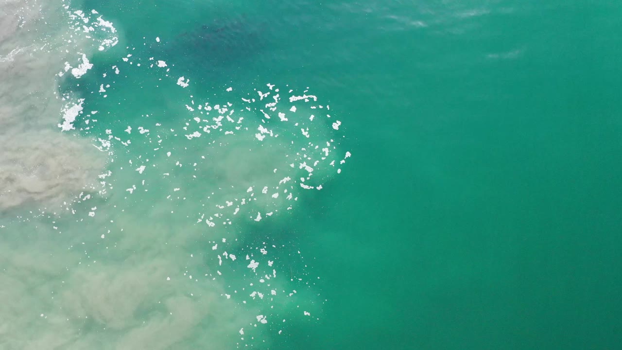 Top-down Los Cabos water view with white sun glints clustered across the middle, cloudy pale surf along the left side, and no visible people.