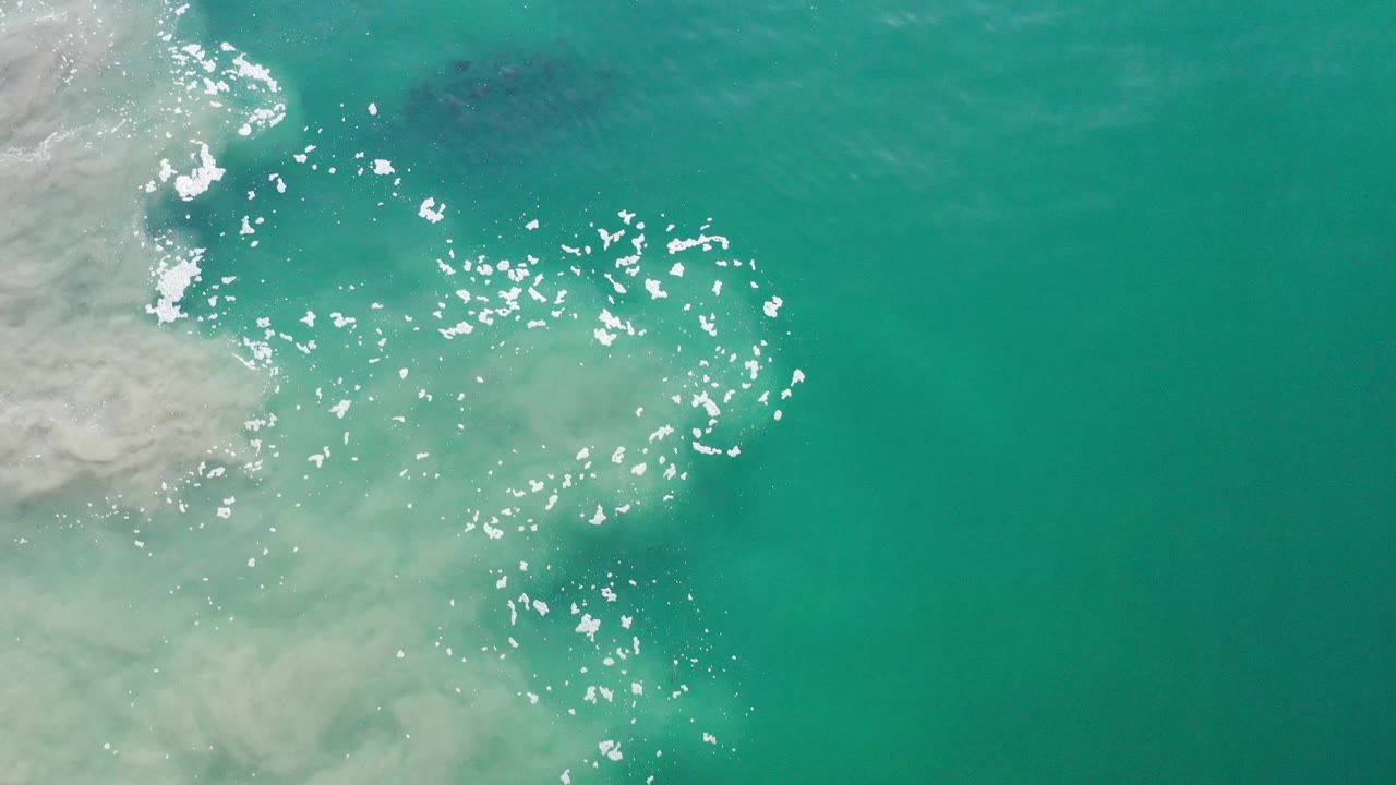 Aerial view over Los Cabos ocean water, with a broad green field of water, bright reflection flecks near the center-left, pale foam at the edge, and no visible people.