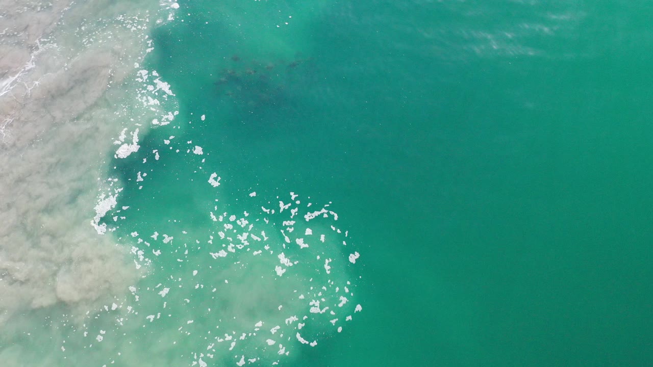 Overhead view of turquoise water off Los Cabos, Mexico, with pale foam entering from the left and no visible people.