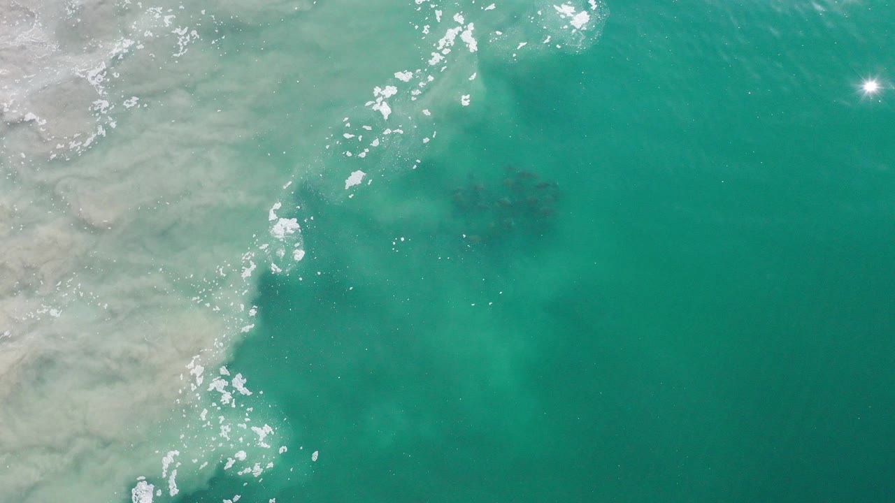 Overhead view in Los Cabos, Mexico, of green-blue water along a pale rocky or sandy edge, with scattered white sun glints and no visible people.