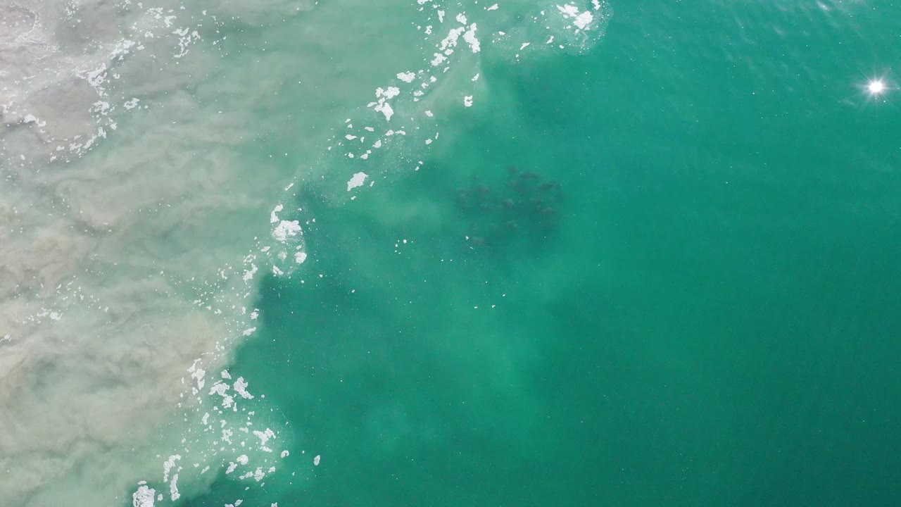 Overhead Los Cabos water frame with the pale edge still visible on the left, bright reflected points near the upper right, and no visible people.