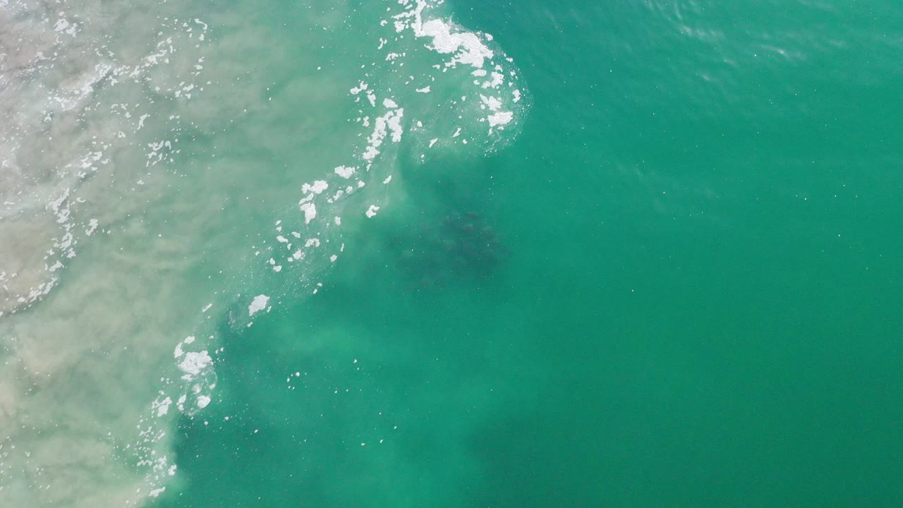 Top-down view of turquoise ocean in Los Cabos, Mexico, with white foam crossing the left side, darker water below the surface, and no visible people.