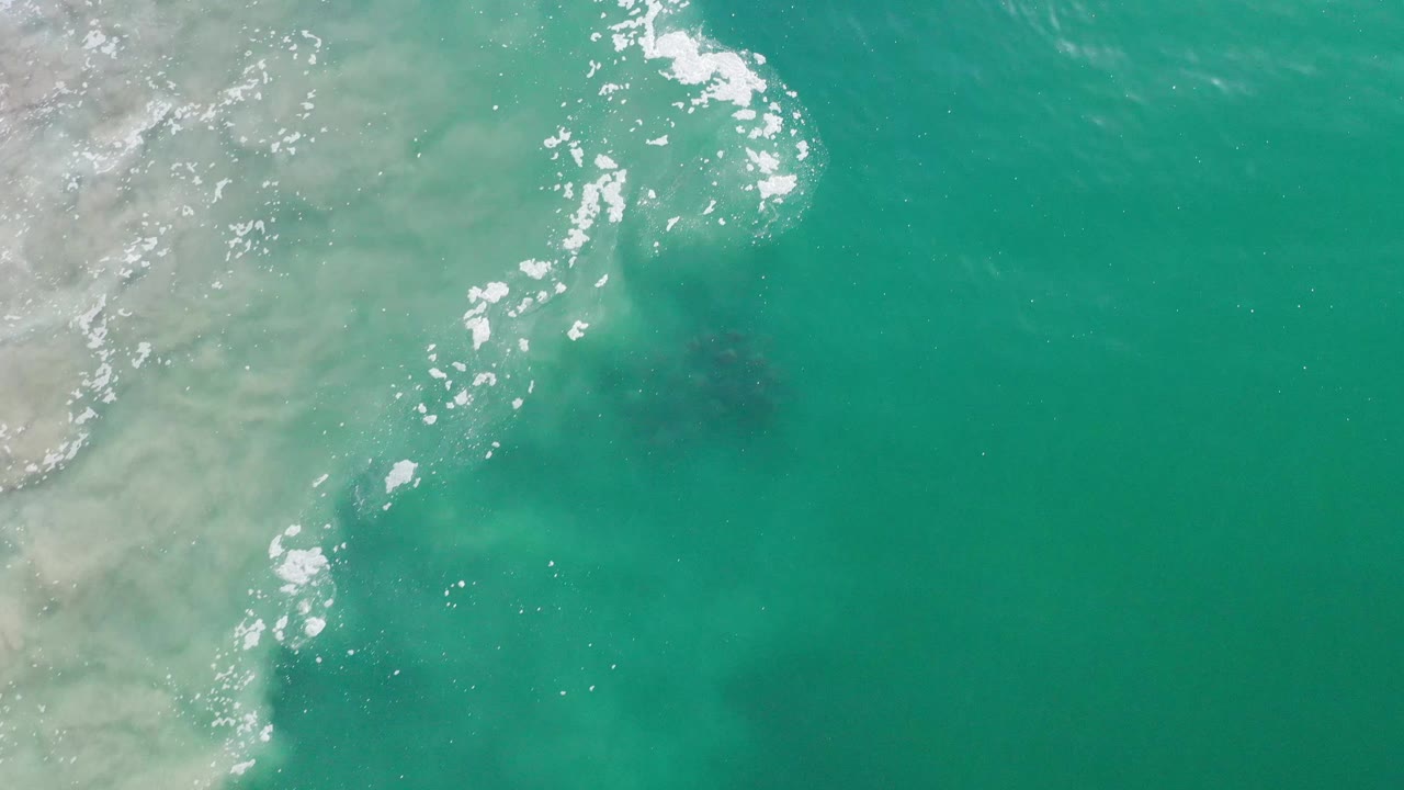 Aerial frame over Los Cabos ocean water with pale suspended sand near the left edge, scattered white foam, and no visible people.