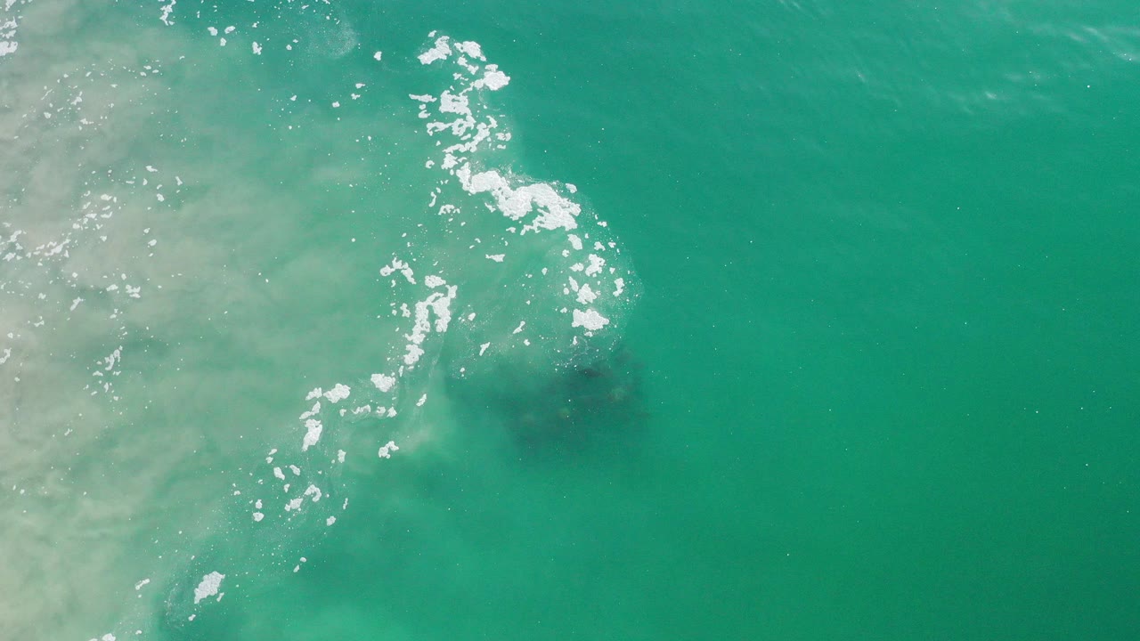 Overhead view of deep green ocean in Los Cabos with white sea foam near the center-left, a pale sand cloud along the left edge, and no visible people.
