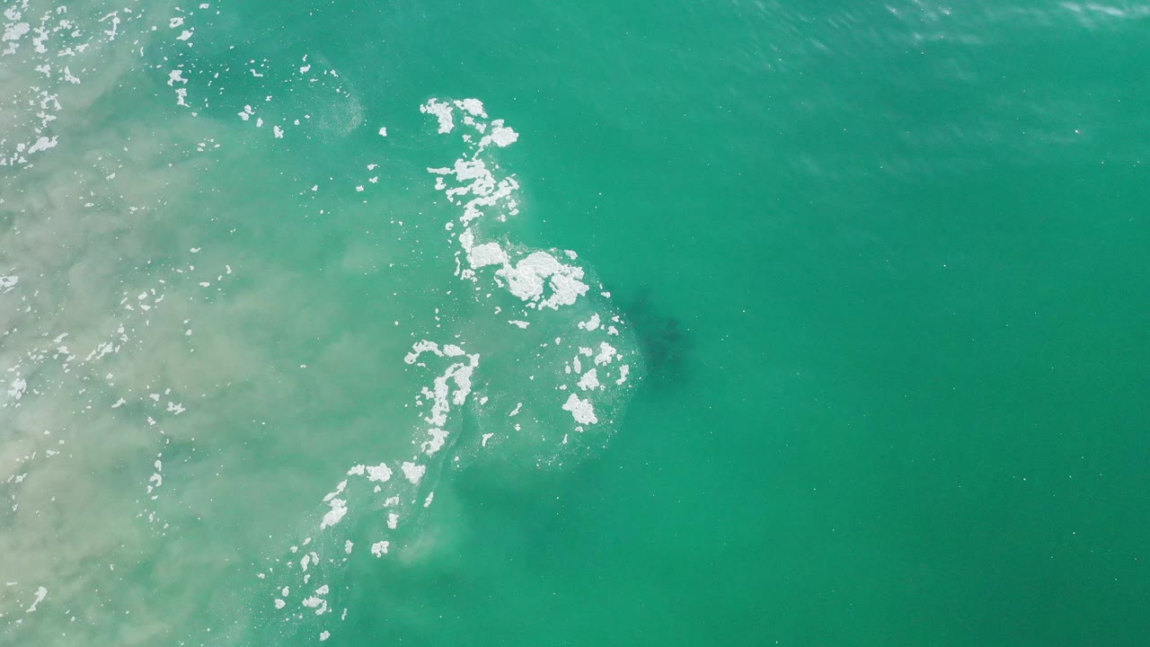 Overhead teal-green water with broken white foam and sand haze.