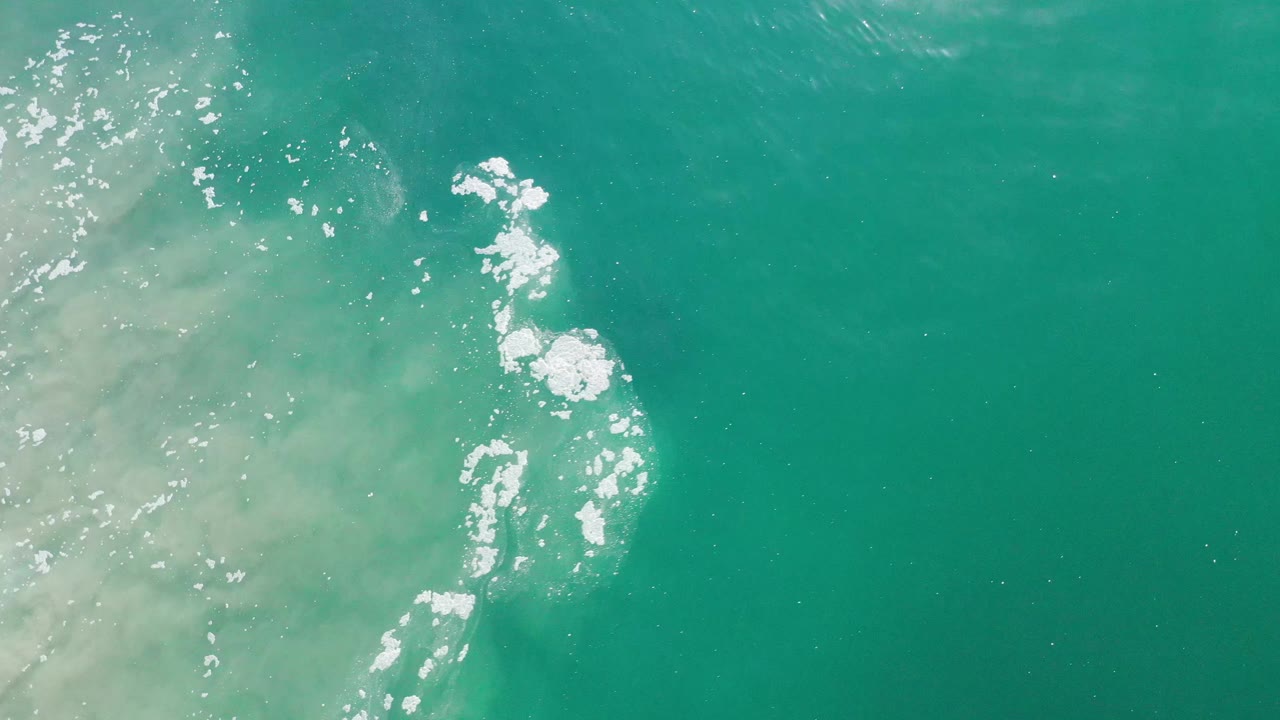 Aerial-looking frame of green-blue water in Los Cabos, Mexico, with bright foam streaks clustered left of center and open water to the right, no visible people.