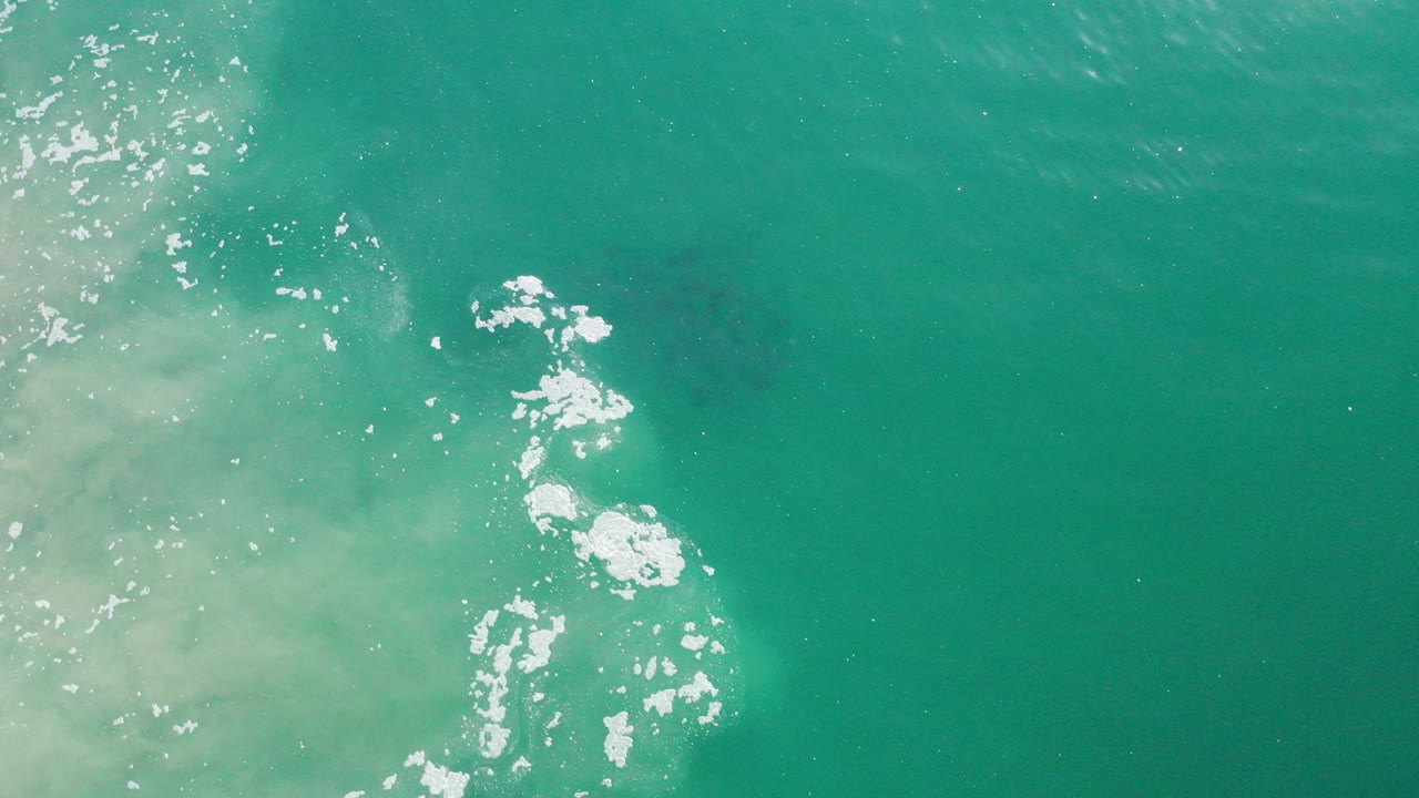 Top-down aerial frame near Los Cabos, Mexico, showing turquoise ocean, pale sandy wash at left, white surface foam near center, and a dark fish school beneath the water with no visible people.