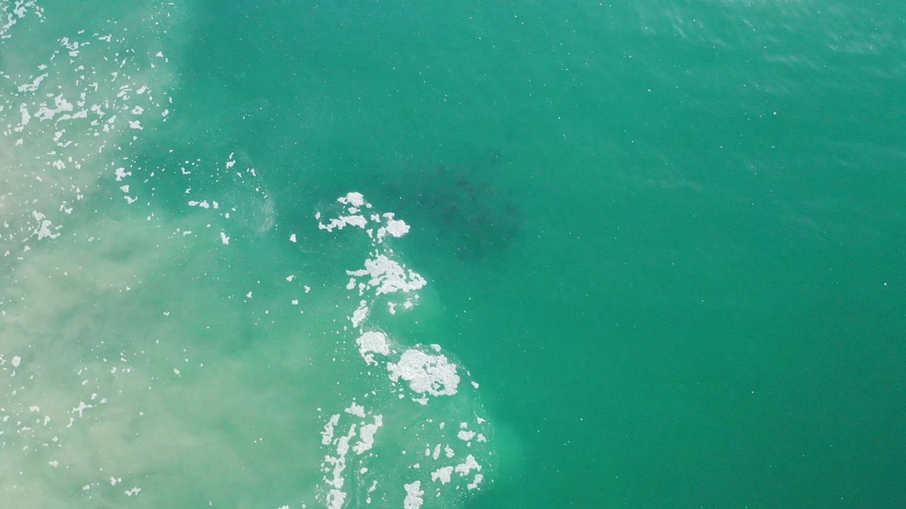 Clear teal ocean with scattered white foam and a darker fish school near the center.