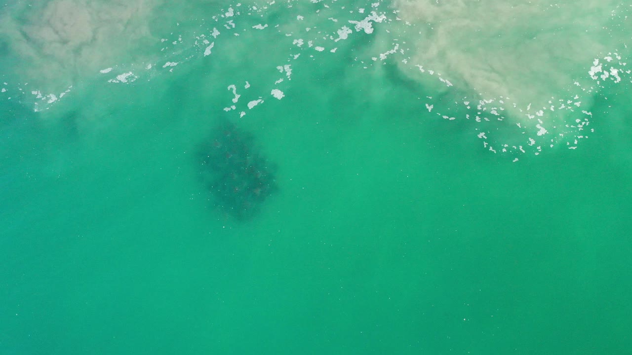 Overhead view of green turquoise sea in Los Cabos, Mexico, with pale wave wash across the upper edge, a dark underwater seaweed patch left of center, and no visible people.