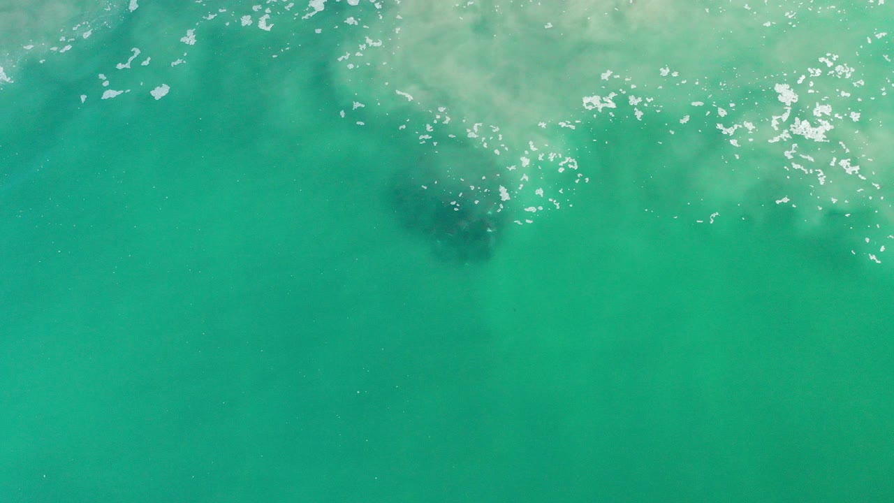 Overhead view of teal ocean water in Los Cabos, Mexico, with pale wave wash along the top edge, scattered sun glints, and no visible people.