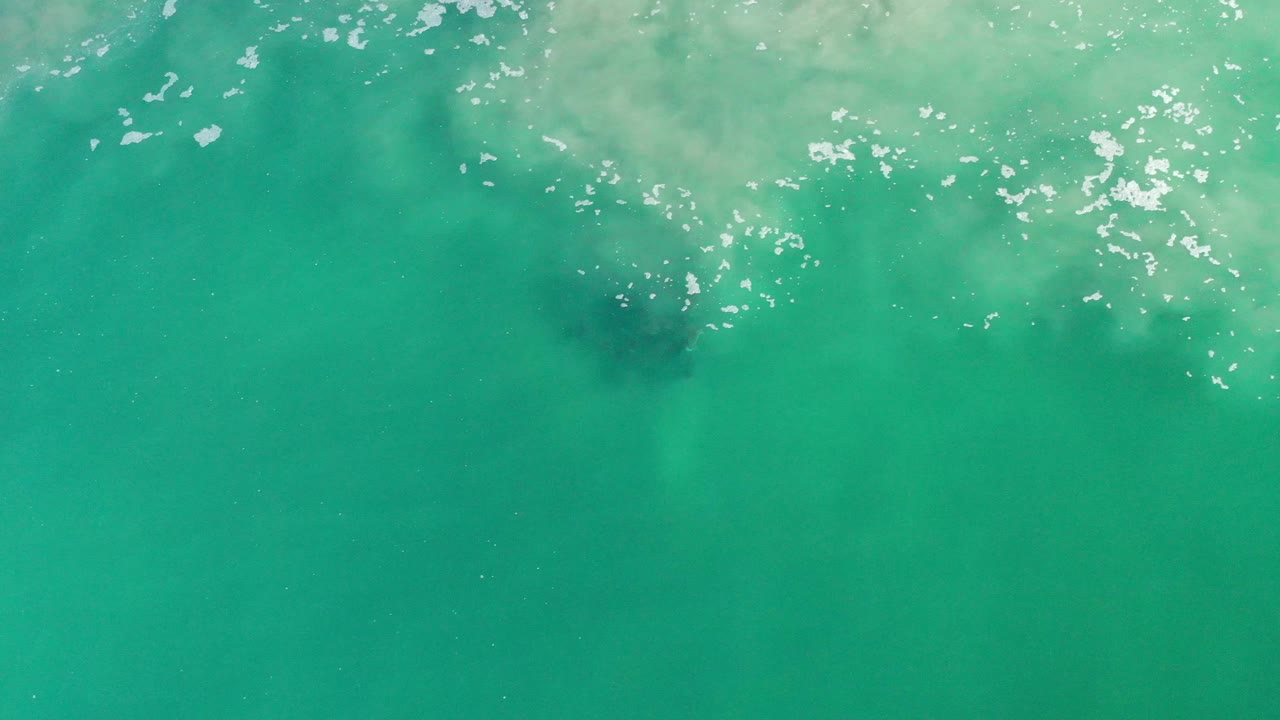 Aerial ocean surface with ripples, glints, and a darker patch below the wash.