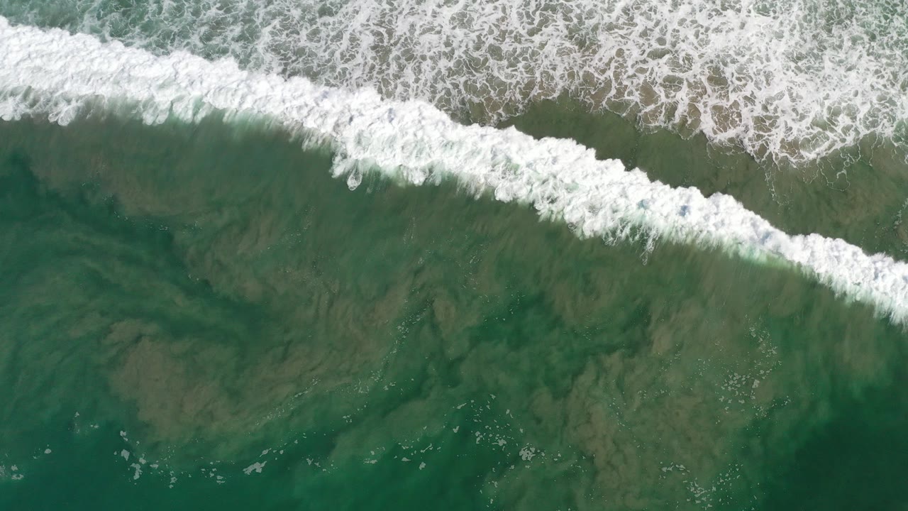 Overhead view in Los Cabos, Mexico showing green ocean water and a narrow white surf band crossing the upper frame, with no visible people.