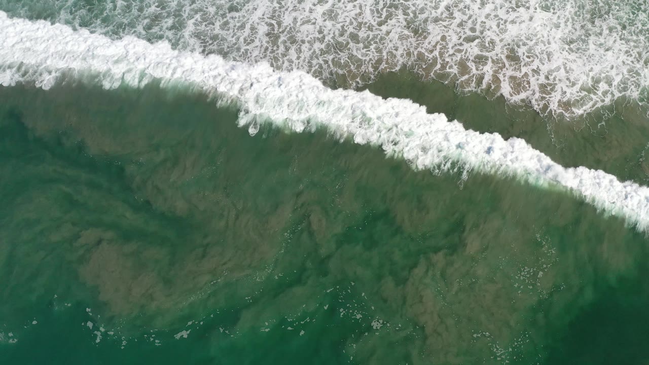 Overhead Los Cabos ocean frame with a thicker white breaking wave across the top third and darker green water below, with no visible people.