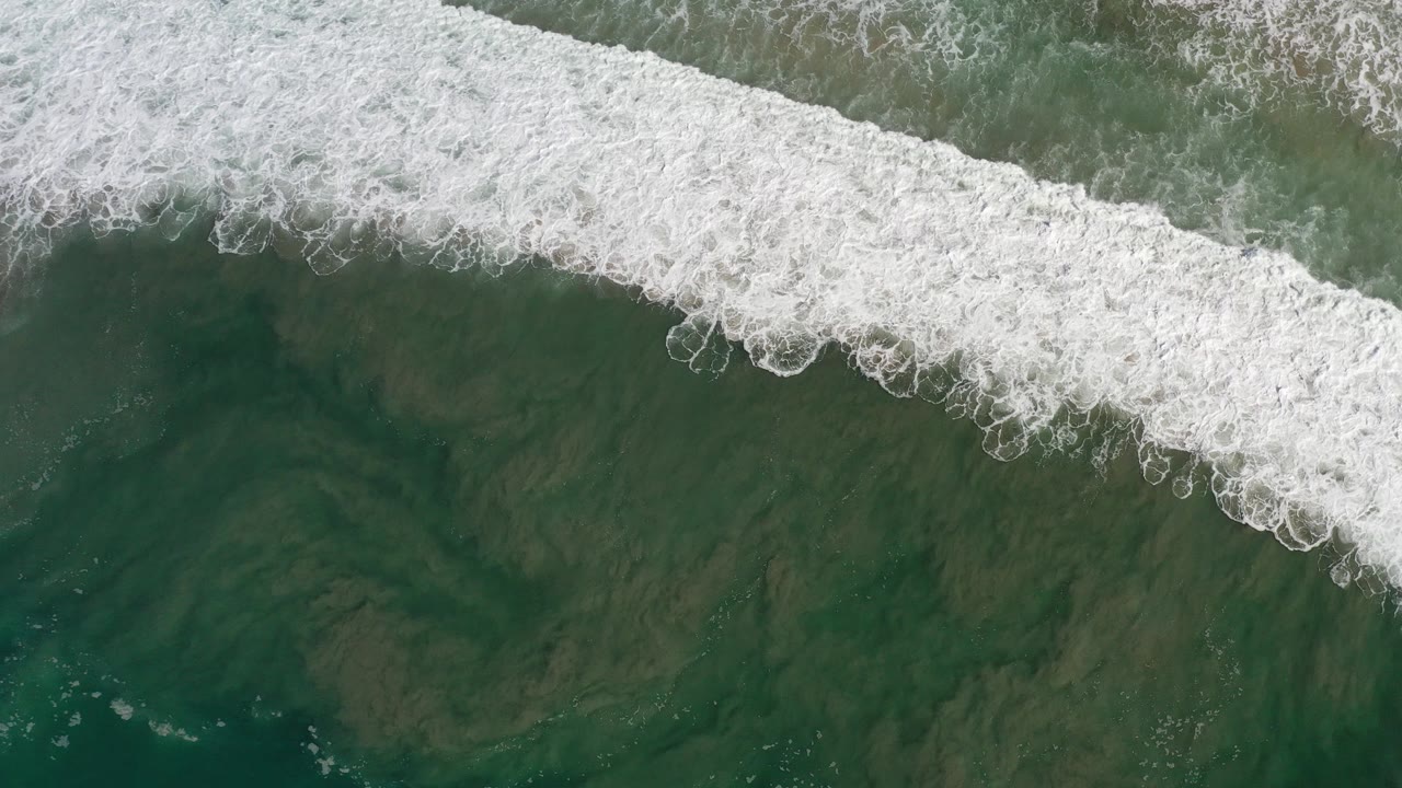 Top-down aerial view in Los Cabos, Mexico, showing a wide band of white foam crossing green water with darker sand patterns below and no visible people.