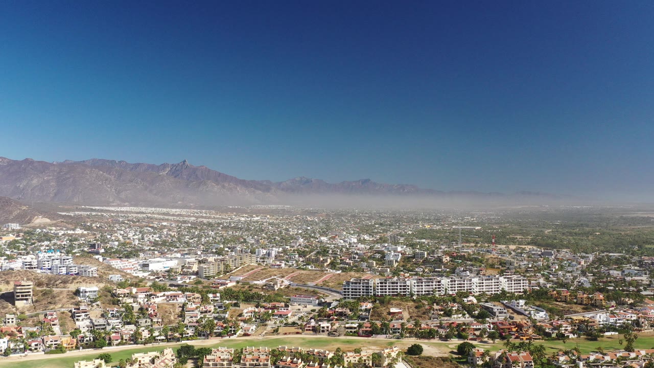 Wide aerial view of a town below mountains under a blue sky.