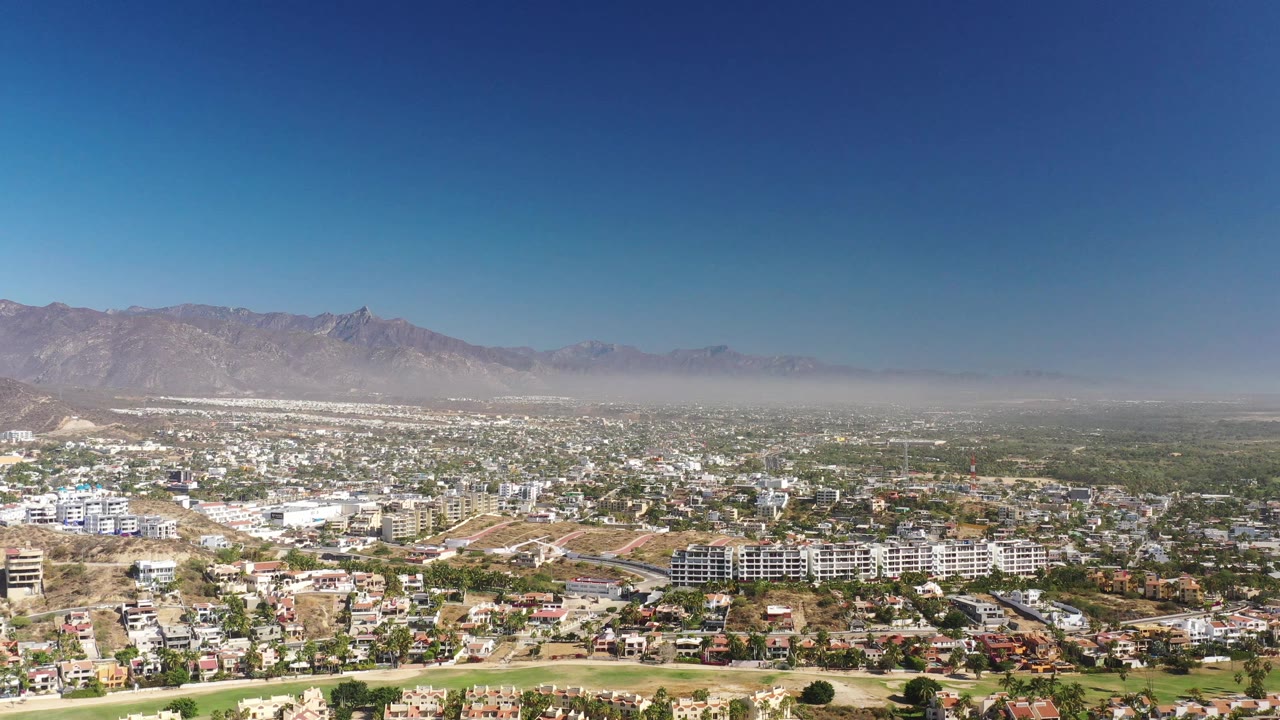 Second frame over Los Cabos shows more of the residential grid and low buildings below the hazy mountain range, with no visible people.