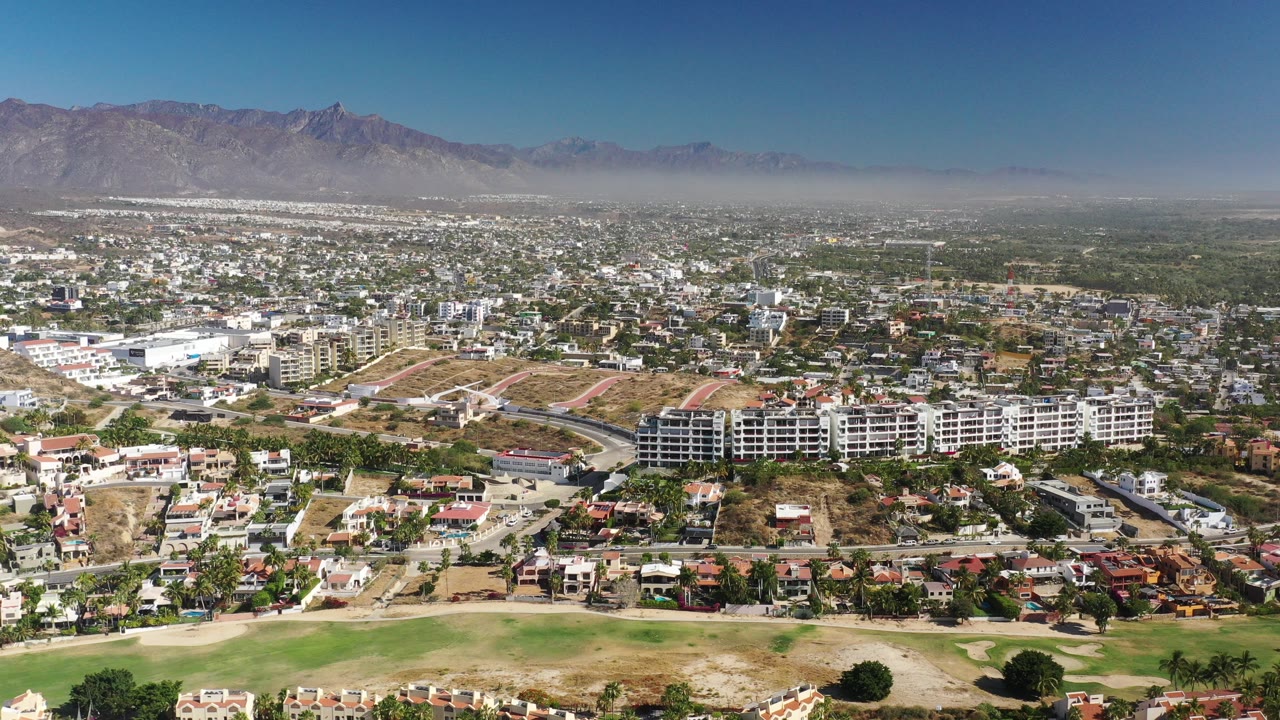 Opening aerial frame over Los Cabos, Mexico, showing green grass in the foreground, residential blocks and construction lots in the midground, hazy mountains in the distance, and no visible people.