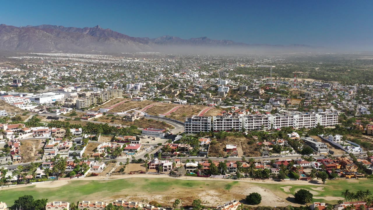 Wide aerial over homes, open lots, apartments, grass, and mountain skyline.