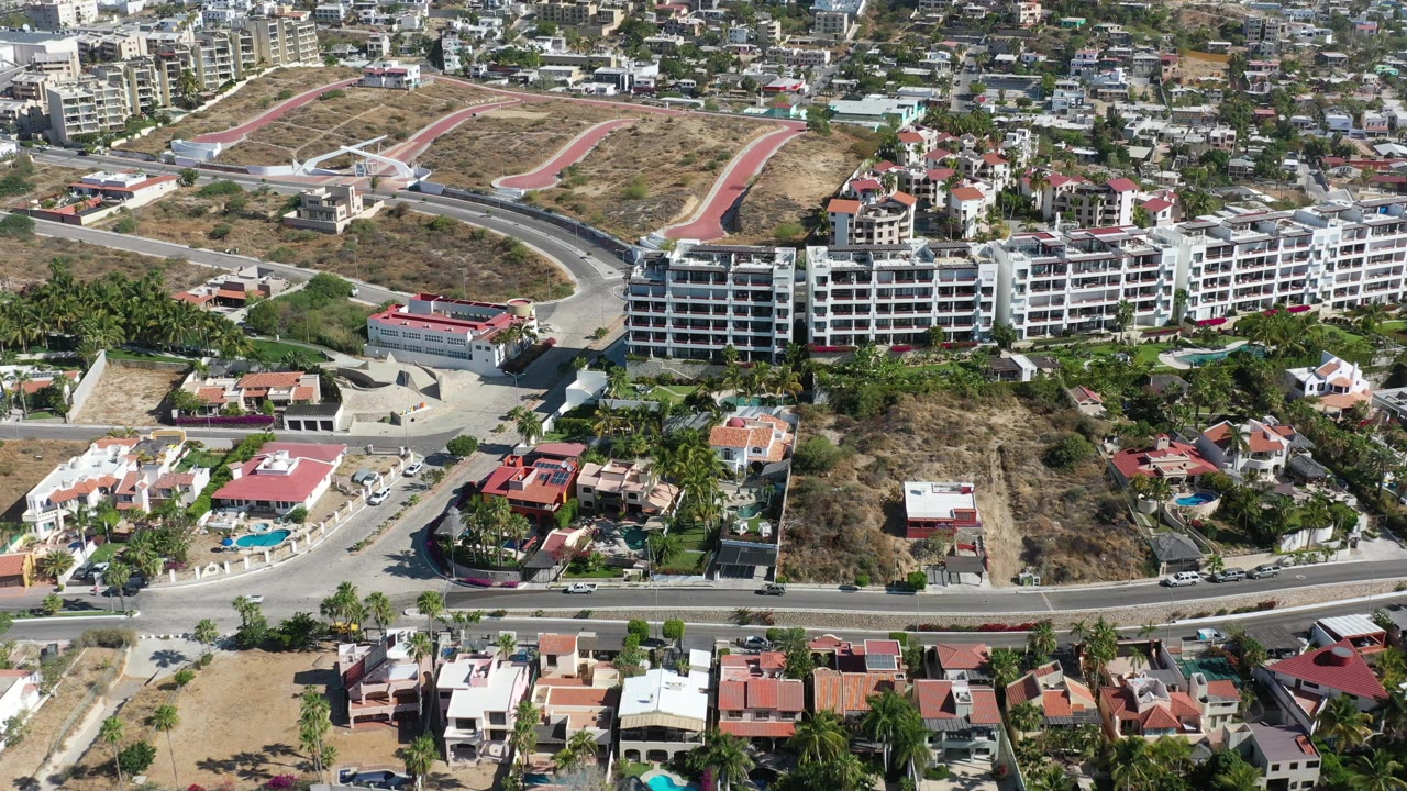 Los Cabos aerial view with the apartment complex centered, a broad curved road below it, dry hillside lots above, and no visible people.
