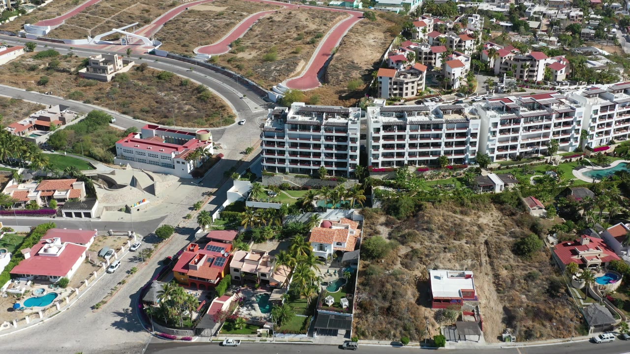 Aerial Los Cabos frame with a long white apartment-style complex, nearby villas, red roofs, parked cars along the roads, dry open land, and no visible people.
